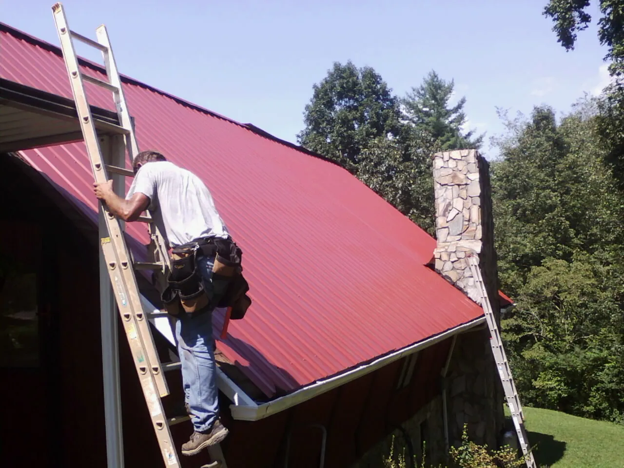 Roofer on a ladder beside a red metal roof and stone chimney — Soffit & Fascia Repair by Roofing Brothers in Woodsfield